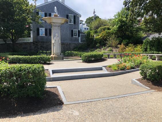 A garden scene featuring a stone fountain with a statue of a woman, surrounded by manicured hedges and a grey house in the background.