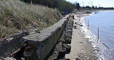 A vertical seawall leaning toward the water with a vegetated slope behind it