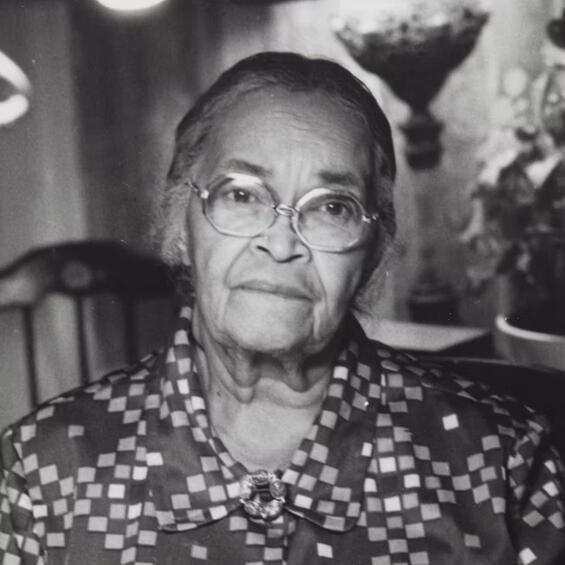 Alt Text: An elderly Melnea Cass sits indoors, with a wooden chair, a lamp, and potted plants visible in the background.