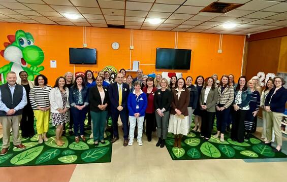 Education Secretary Steve Zrike stands with a group of local and state officials in an early child hood program in Pittsfield. Playful green children's rugs cover the tiled floor and a large cartoon Yoshi dragon is painted on the orange wall behind the group. 