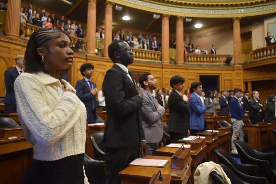 Students stand in the Massachusetts House Chamber during Student Government Day, facing forward with their right hands over their hearts as if during the Pledge of Allegiance. The foreground shows a group of formally dressed high school students—one in a light sweater and others in suits—while additional participants and observers fill the chamber floor and balcony behind them.