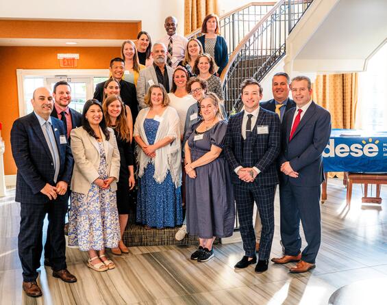 A group of state officials, including Secretary Zrike and Commissioner Martinez stand on a stairwell in business attire. A blue sign with the DESE logo is behind them.