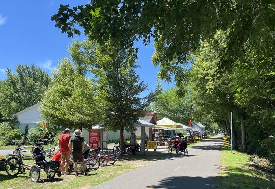 A variety of adaptive cycles and popup tents line a paved bike trail as people are fitted for cycling.