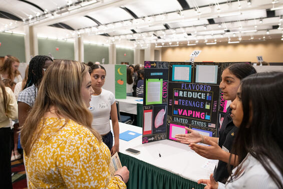 A group of female students speak to each other in front of civics project showcase display. A large room with other displays is visible behind them. 