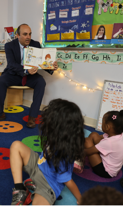 Secretary Zrike sits on a chair holding a children's book out so that children seated on a carpet in front of him can see the images in the book. Classroom decorations hang on the walls beside them.