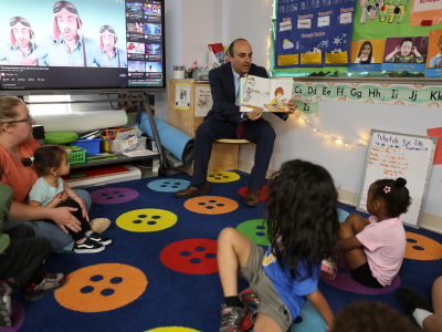 Secretary Zrike sits on a chair holding a children's book out so that children seated on a carpet in front of him can see the images in the book. Classroom decorations hang on the walls beside them.