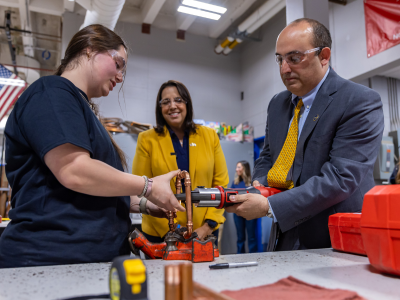 Secretary Zrike in a suit is holding a tool and a student is helping him work on a piece of metal in a vice grip in a high school lab. Lieutenant Governor Kim Driscoll in a yellow suit is smiling and watching in the background.