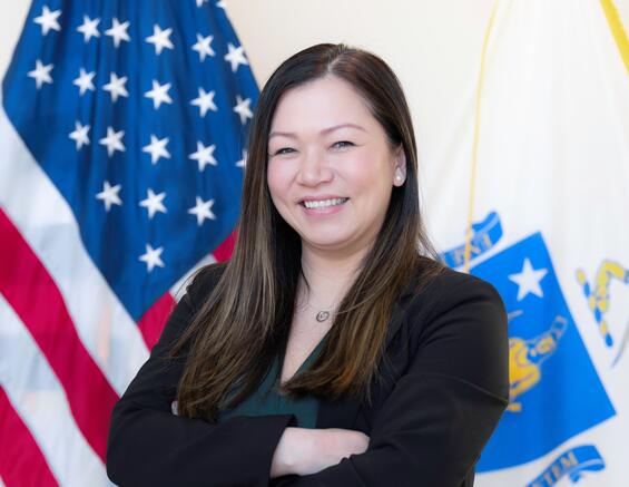 Truc DeCoste smiling in front of the USA and Massachusetts flag