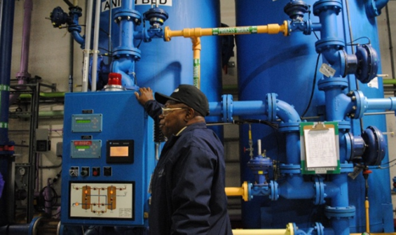A worker stands beside industrial manufacturing equipment and a control panel in a production facility, with tanks and piping visible in the background.