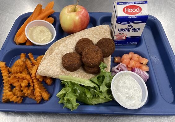 A blue tray with milk, an apple, french fries, carrot sticks and vegetables sits on a gray lunch table.