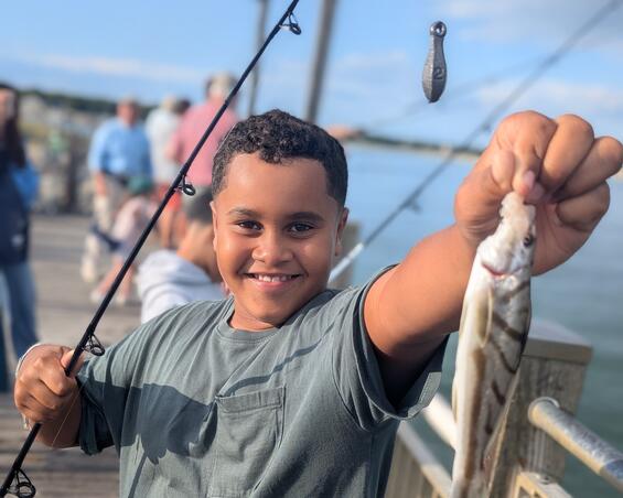 A smiling boy holds up the fish he caught on an ocean pier.