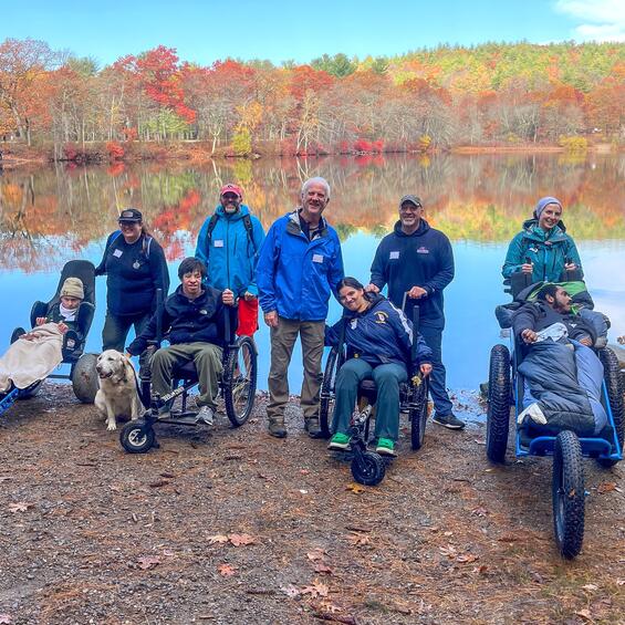 A group of participants using a variety of all terrain wheelchairs pose for a picture along the shore of a lake in autumn.
