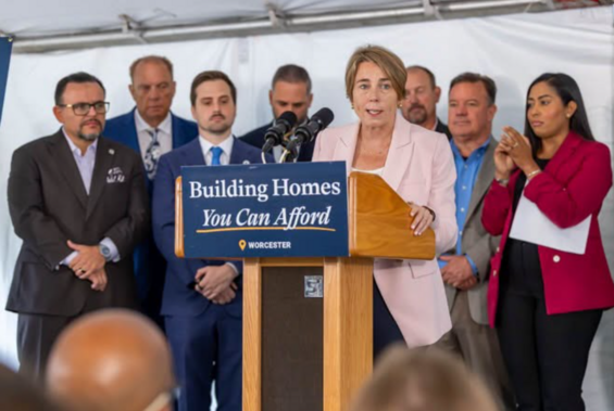 Governor Healey stands at a podium with several other officials behind her. The podium has a sign on it that reads "Building homes you can afford."