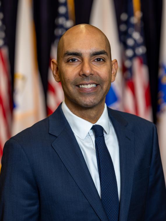 Secretary Jon Santiago Headshot in front of the Commonwealth of Massachusetts and United States of America flags.