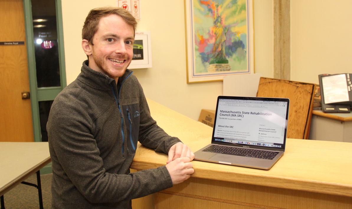 White man with red hair standing next to a laptop