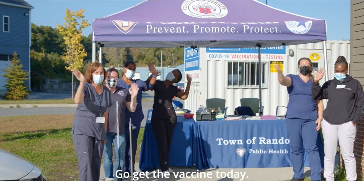 Group of people and healthcare works waving in front of vaccination clinic tent with "Town of Randolph" branding and a caption: "Get the vaccine today."