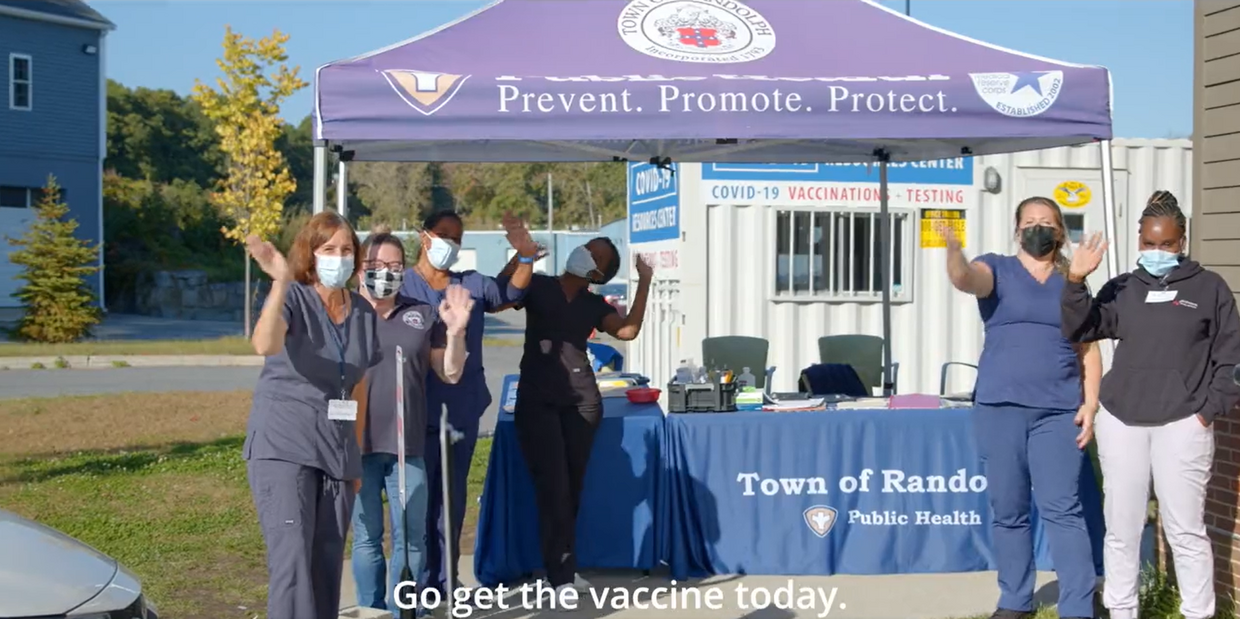 Group of people and healthcare works waving in front of vaccination clinic tent with "Town of Randolph" branding and a caption: "Get the vaccine today."