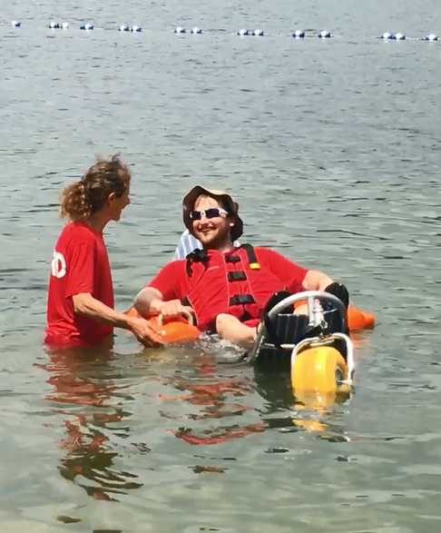 A man is floating in the water in a floating beach wheelchair. He is wearing a life jacket and resting against the fabric backrest of the chair. A woman is standing beside him, with her hands on the frame of the chair.
