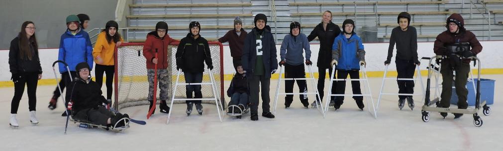 A large group of young adult skaters is on the ice in front of a hockey net. Some skaters are using skate walkers, some skaters are using ice sleds, and one skater is using a walker with a harness.