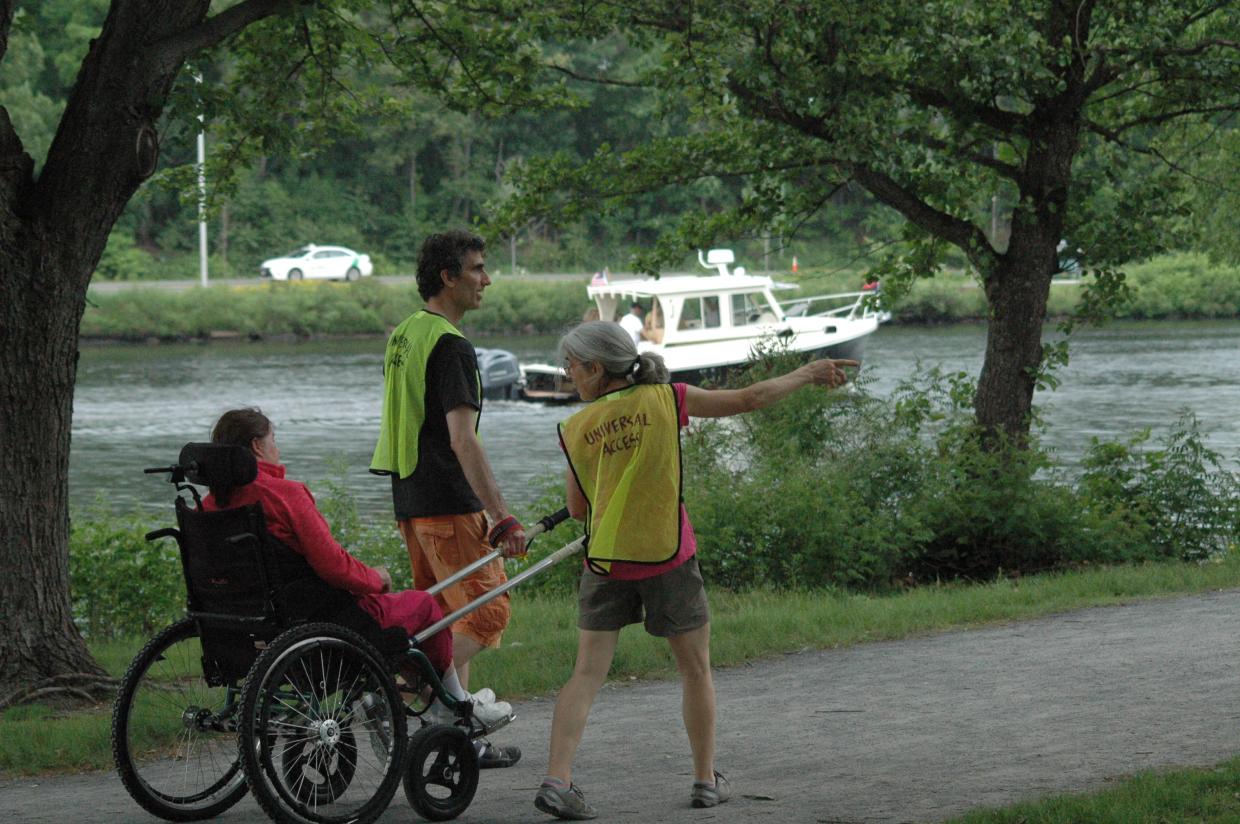 A person in a hiking wheelchair is being pulled by two people in Universal Access vests. The people pulling the hiking wheelchair are using long poles. The group is walking along a trail by a river. Trees line the riverbank.