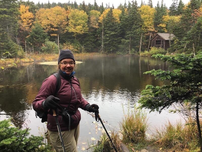 A woman stands in front of a pond, holding hiking poles.