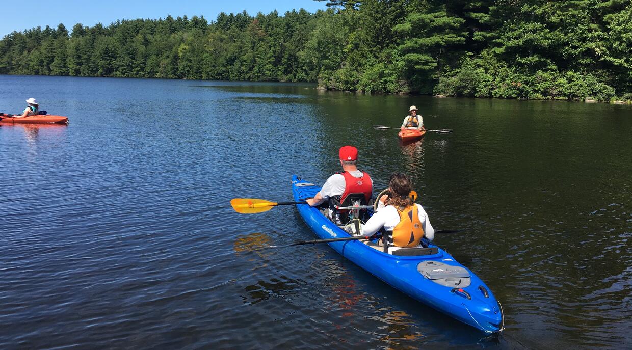 Two people are pictured in a blue tandem kayak leaving shore at Highland Lake. Two other people are in the distance in single kayaks.
