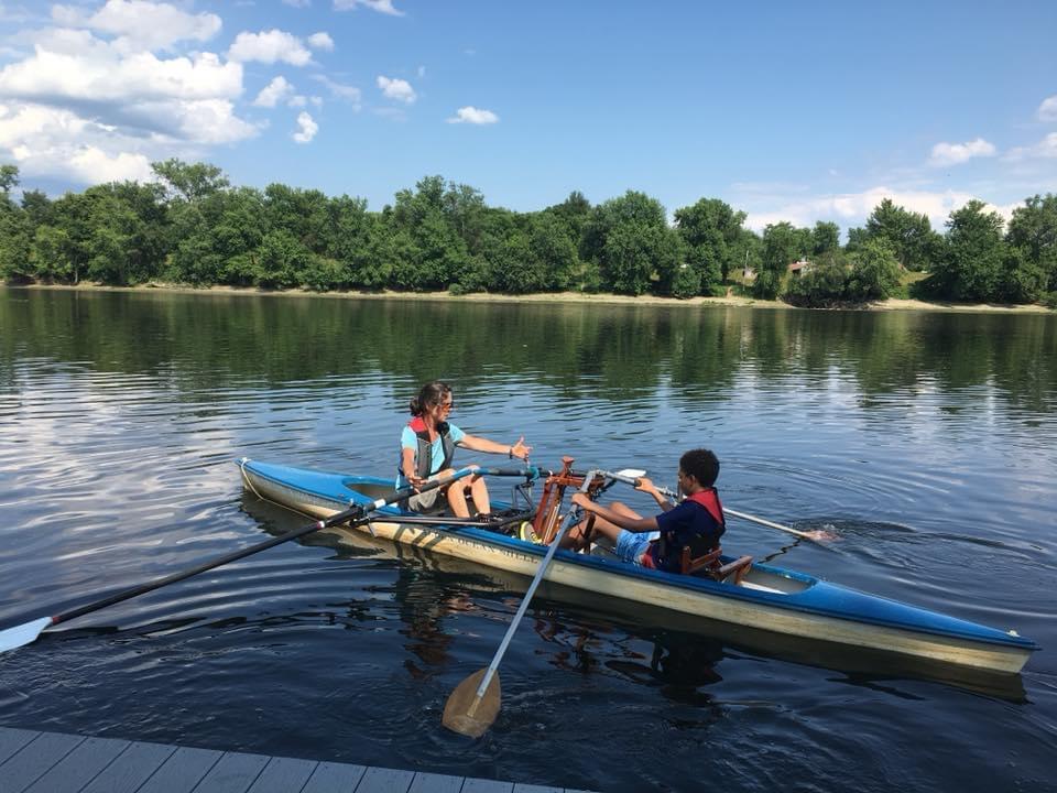 Two people are seated facing each other in a Front Rower, a rowing system which allows you to row with one side of the body. Trees hover over the river in the distance.