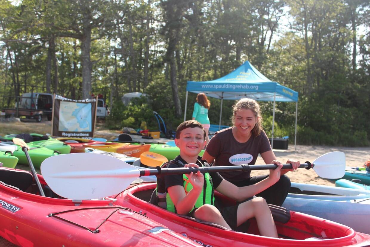 A teenager is seated in a red kayak holding up a paddle and smiling at the camera. An instructor is looking at him smiling. There is a blue tent and brightly colored kayaks visible on shore behind them.