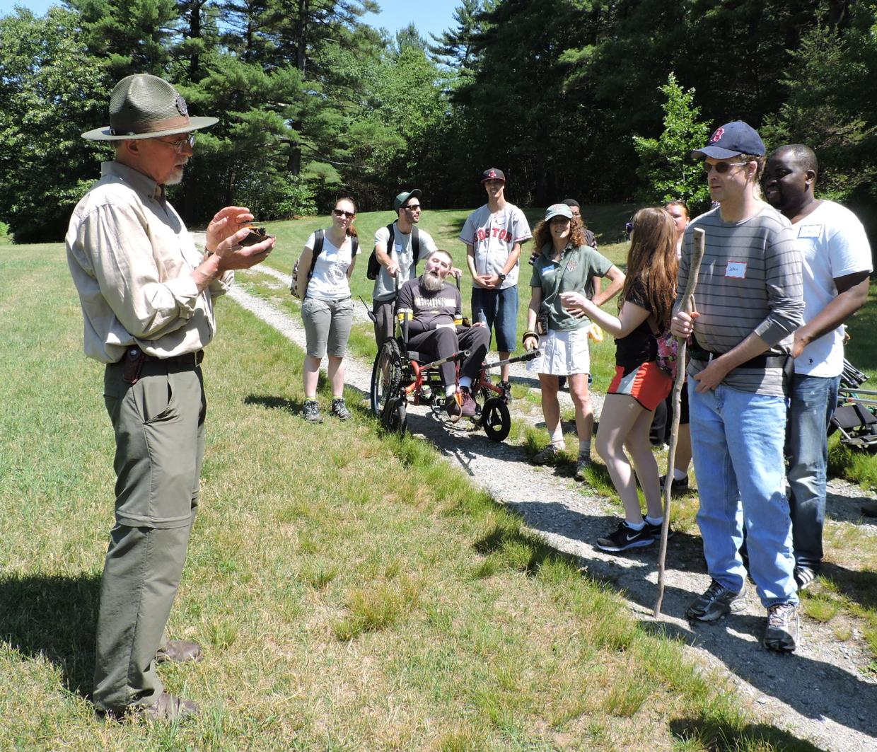A group of hikers listens to a DCR staff member holding a turtle. Some of the hikers are using hiking wheelchair and some are using walking sticks.