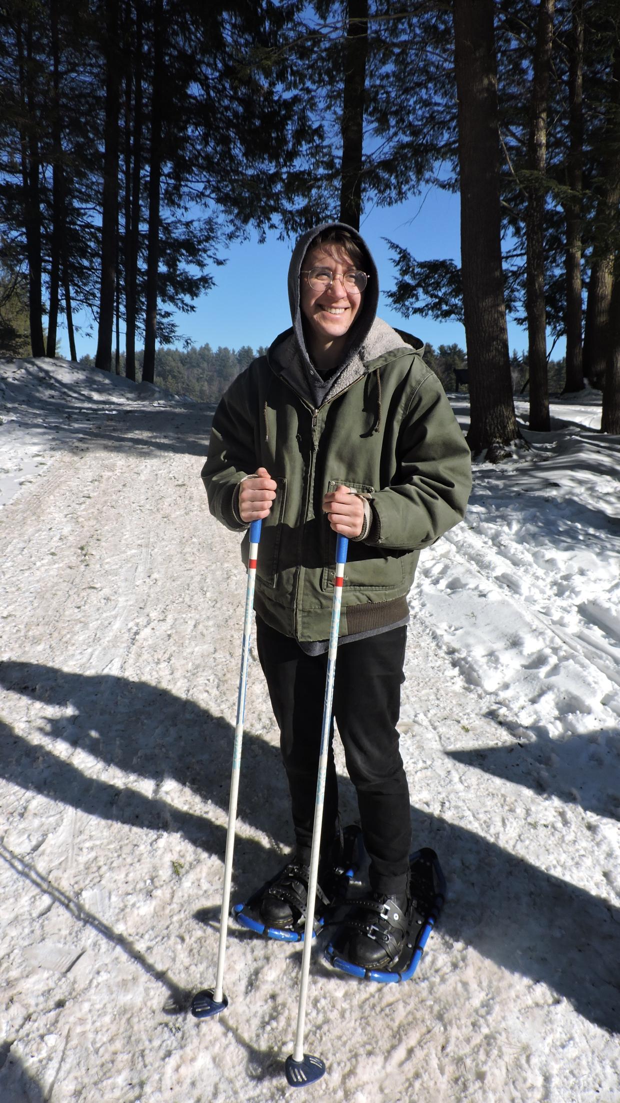 A person wearing snowshoes and holding ski poles on a plowed, snowy, wide, path in the woods.