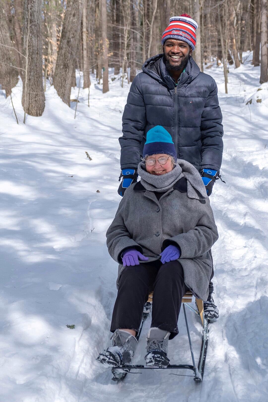 Two people smiling on a snowy trail in the woods. One is sitting on a sled, the other standing behind her.