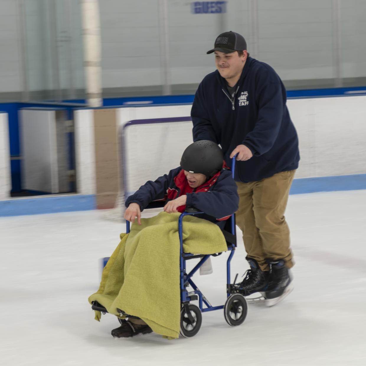 A person on skates pushes a person in wheelchair on ice at an ice skating rink.