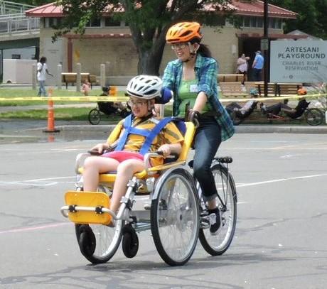 Two people riding an adaptive tandem bicycle in a closed parking lot. A sign reading Artesani playground in the background. 