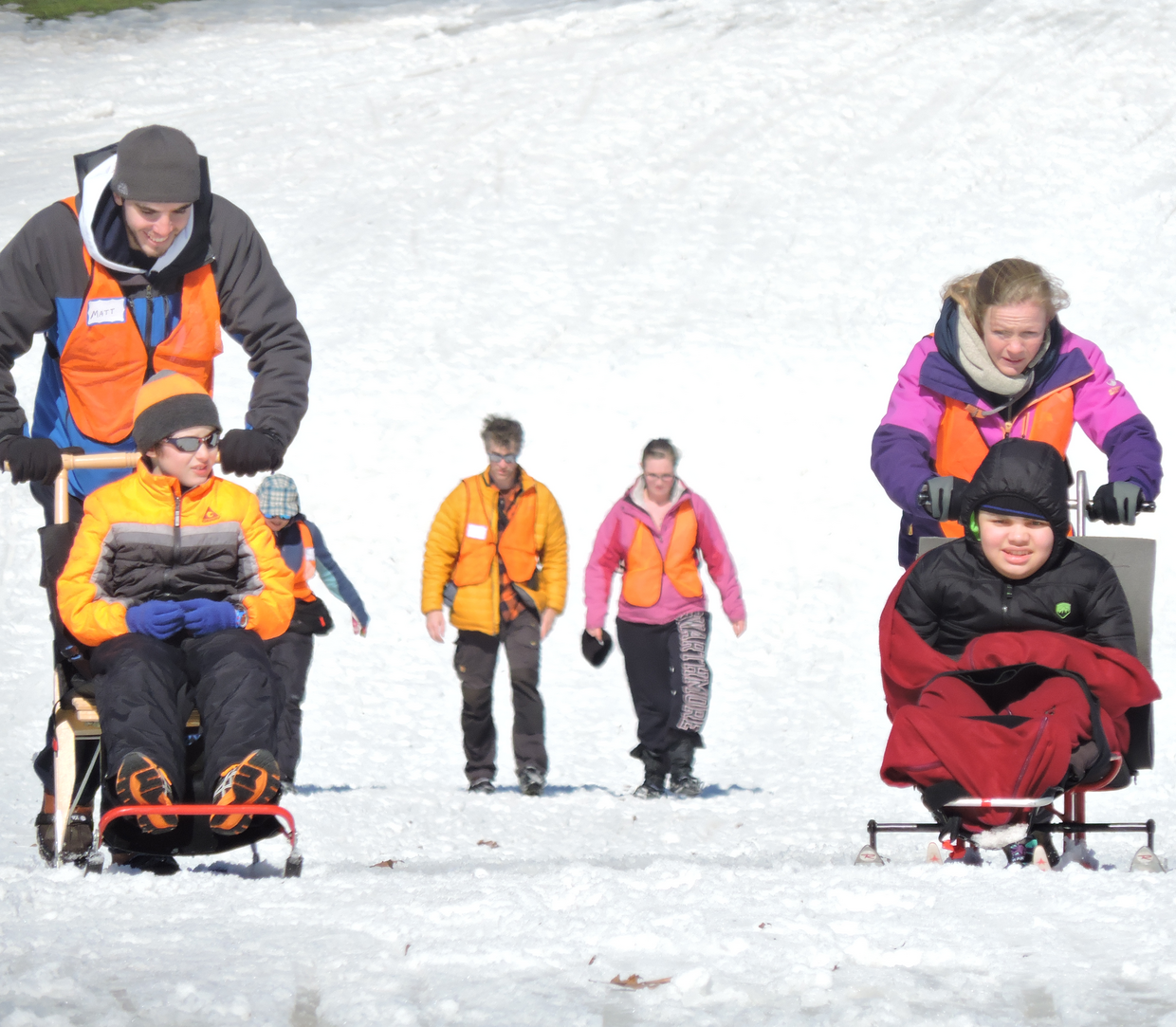 A group of people, smiling while skiing down a snowy hill. Two using sit skis while being pushed from behind.