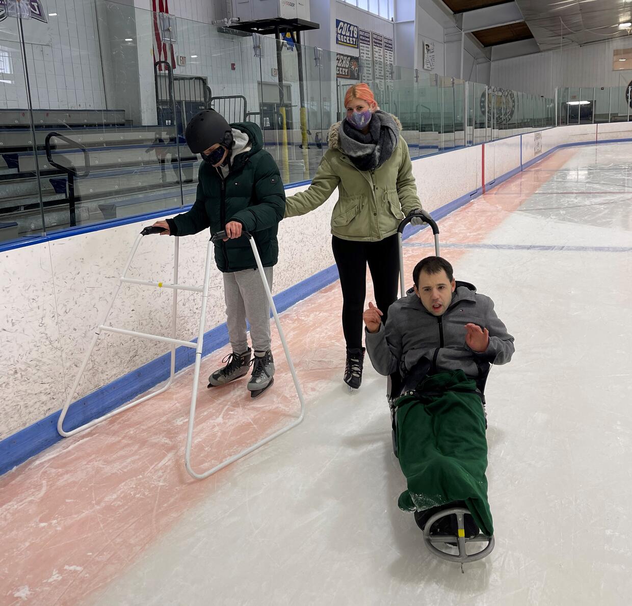 Three people ice skating at an indoor rink using an ice sled, ice skates, and a skate walker. 