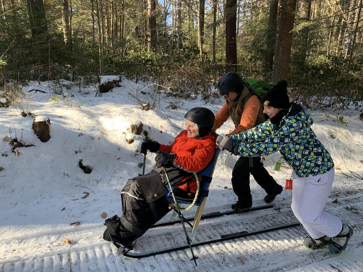 A woman skiing on a kick sled while two others push from behind. 