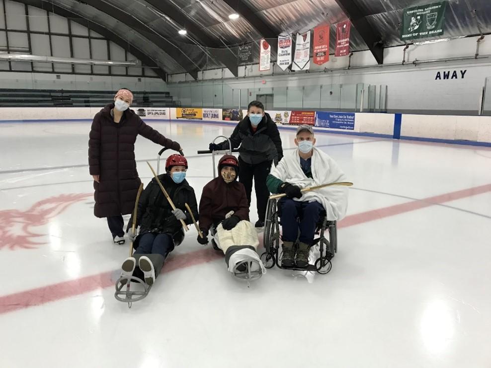 5 skaters on the ice at an indoor rink in a group looking at the camera. Two skaters are in sleds, on is sitting in a wheelchair. Three of the skaters are holding adaptive hockey sticks.