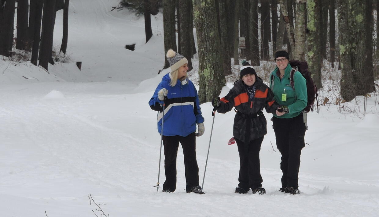 A picture of three smiling people standing in the snow in winter clothes with trees behind them. Two are holding hiking poles. 