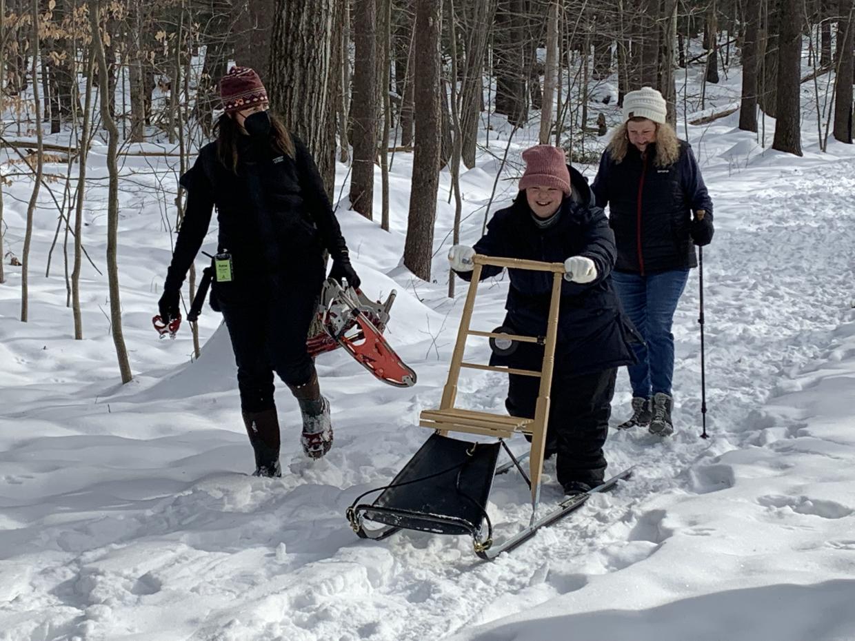 Three people walking on a snowy trail in the wood. One is pushing a kick sled, one is holding snow shoes and one is walking with trekking poles. 