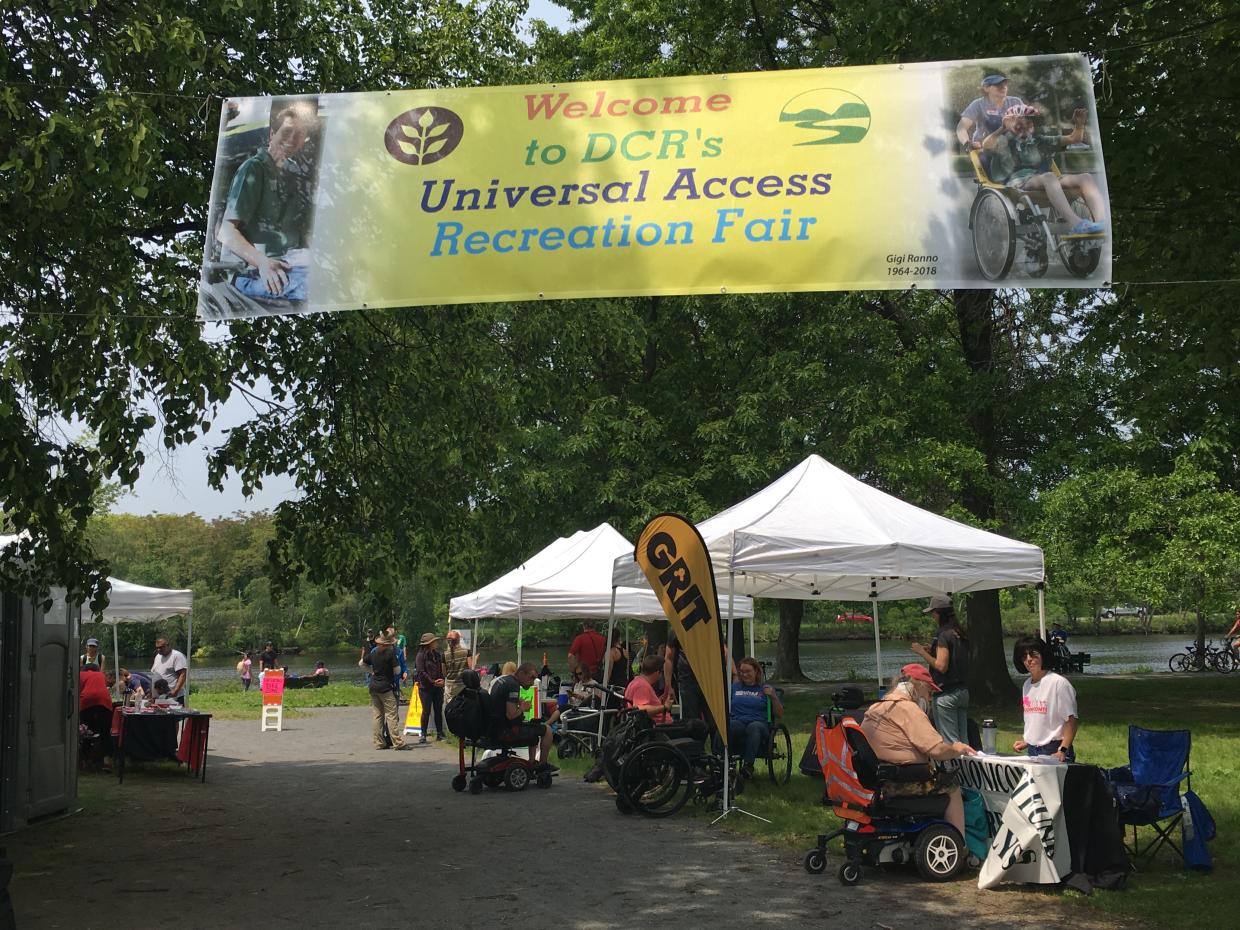 A banner reading "Welcome to DCR's Universal Access Recreation Fair" hangs over a shaded stone dust path. Tents with tables are set up along the path edge. People are gathered at the tents talking to each other.  
