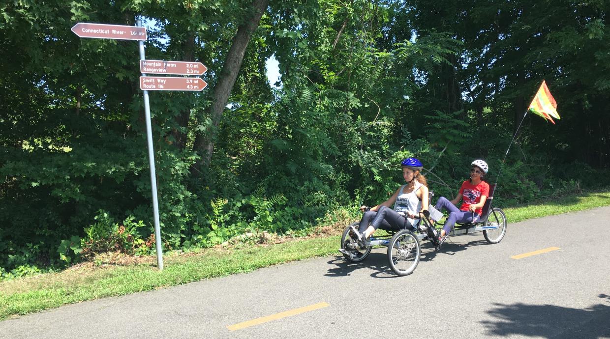 Two rider on a recumbent tandem tricycle peddle on a bike path past some trees. A sign points left to the Connecticut River and right to  Mountain Farms, Rangeview, Swift Way, and Route 116.