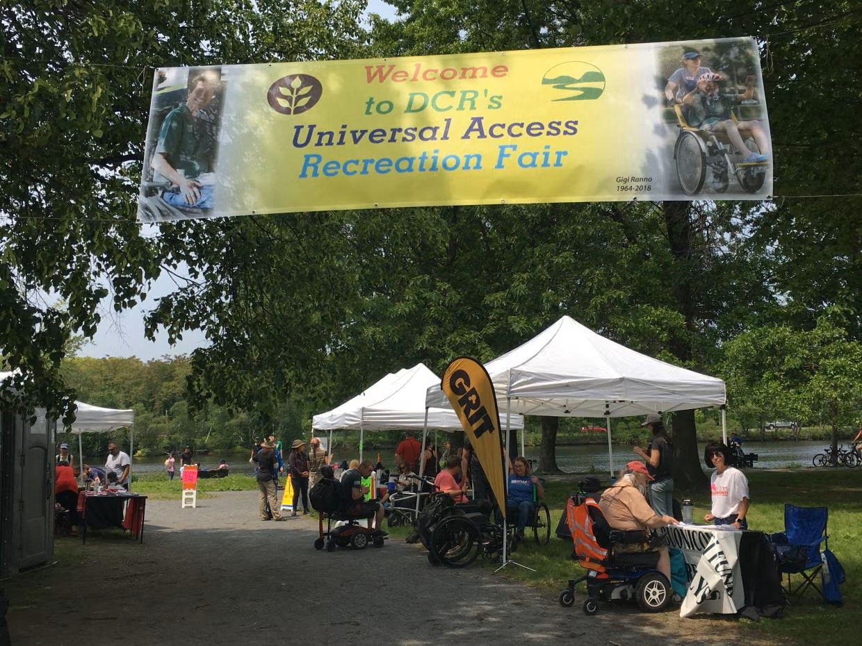 A banner reading "Welcome to DCR's Universal Access Recreation Fair" hangs over a shaded stone dust path. Tents with tables are set up along the path edge. People are gathered at the tents talking to each other. 