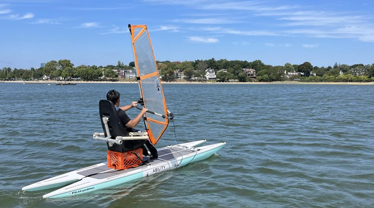 A windsurfer is surfing while sitting on a crate in a seat with back support.