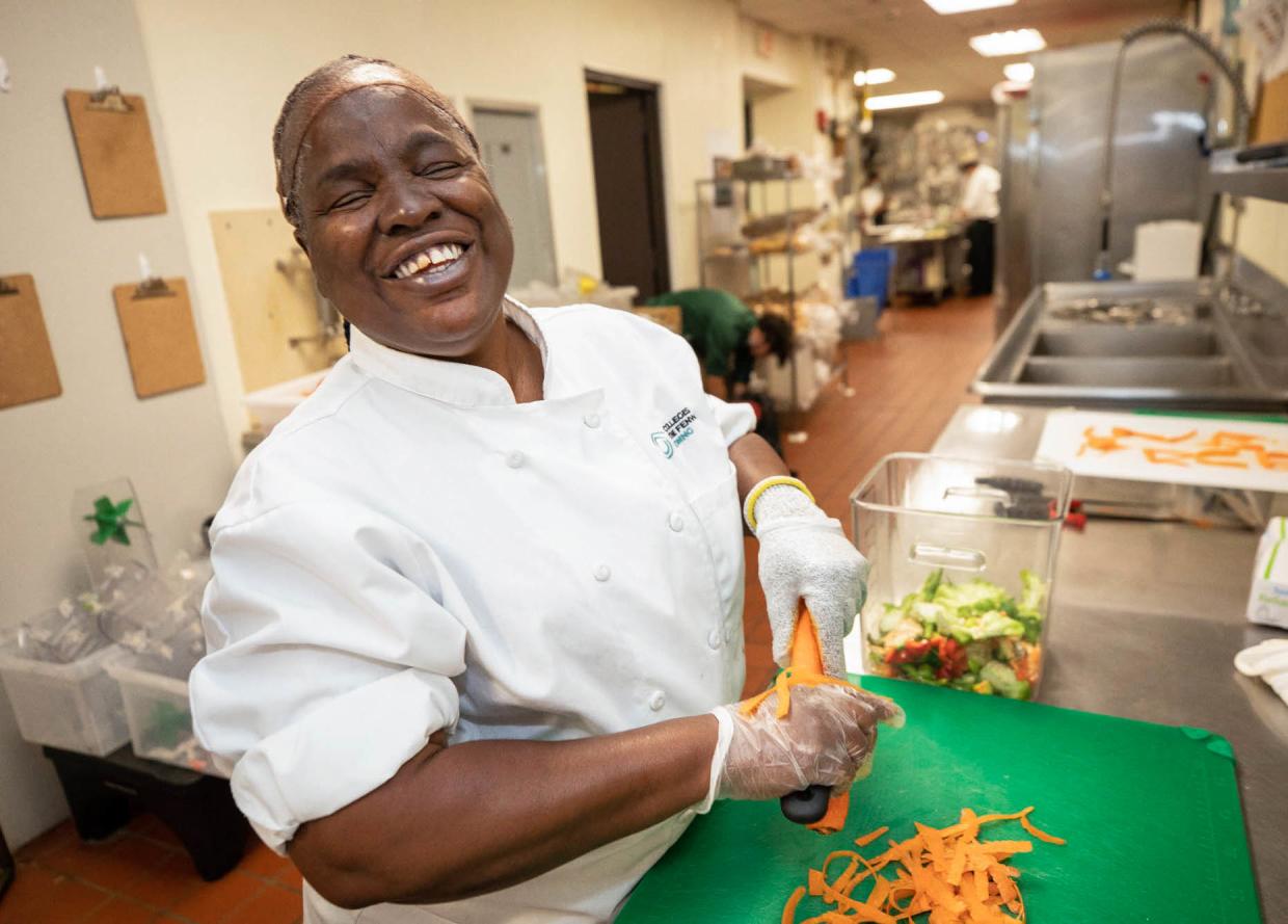 A photo of a woman smiling while in a commercial kitchen cutting a carrot