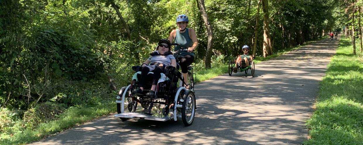 Two cyclists share a tandem. The cyclist in front is sitting in a wheelchair that is on a platform, while the person behind pedals. Another cyclist follows on a recumbent tricycle.
