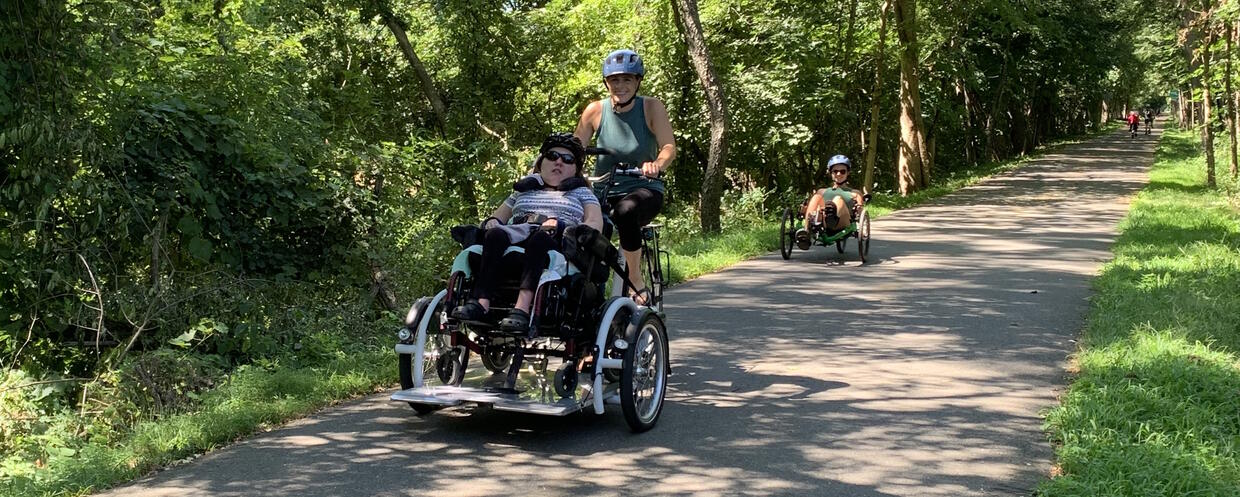 Tandem cyclists using a wheelchair cycle on a paved bike path.
