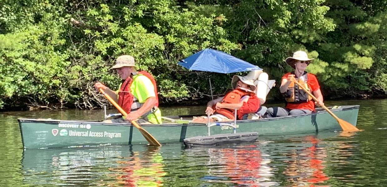 Three people in a canoe with outriggers. The person in the middle is using a seat with foam supports and an umbrella for shade.