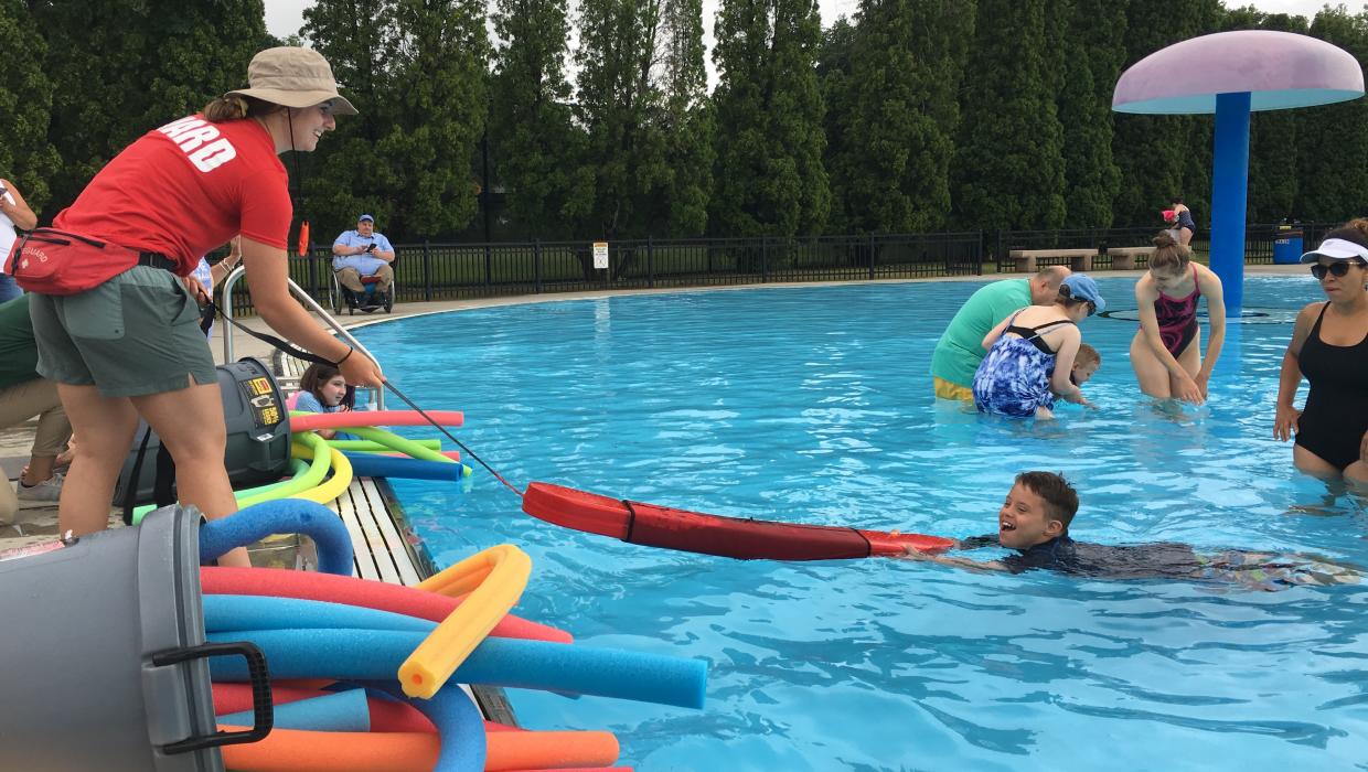 a lifeguard tows a child holding onto a float in a pool. There are buckets of colorful pool noodles at the edge of the pool.