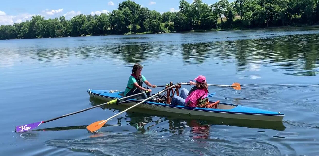 Two people share a sculling shell. The people are facing each other. The person facing forward is rowing with one arm on an oar, and another oar attached to a frame in front of them.