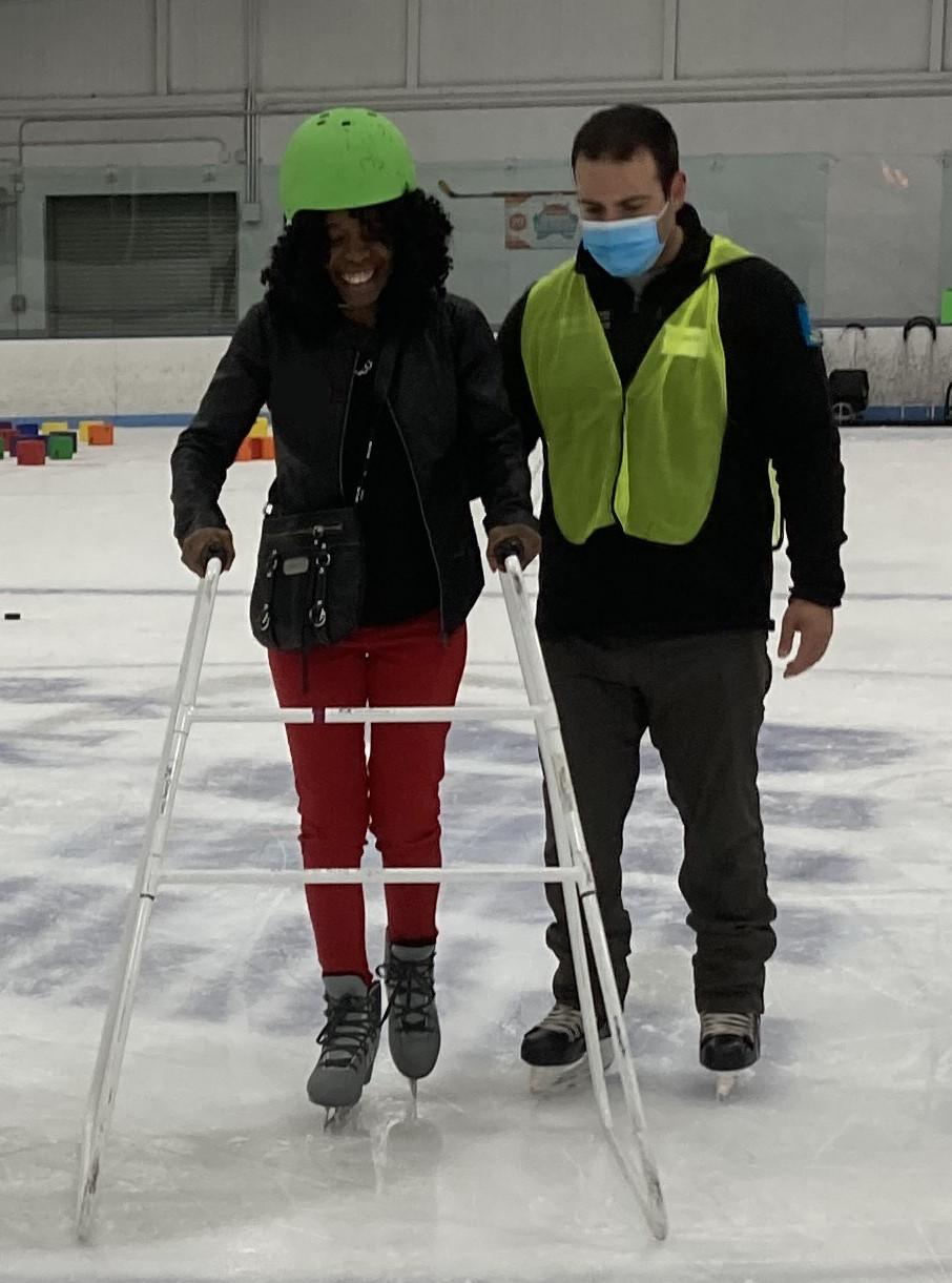 A skater wearing skates and a helmet is pushing a walker in front of them. A staff person is skating beside them with a hand on their back.